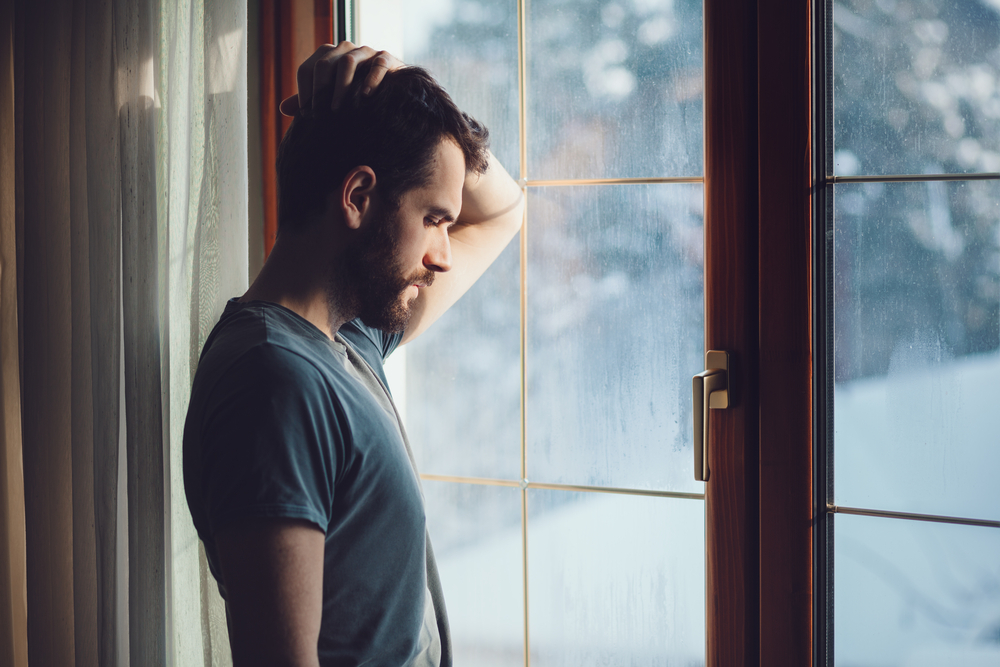 Man in contemplation looking out a window with a hand on his head.