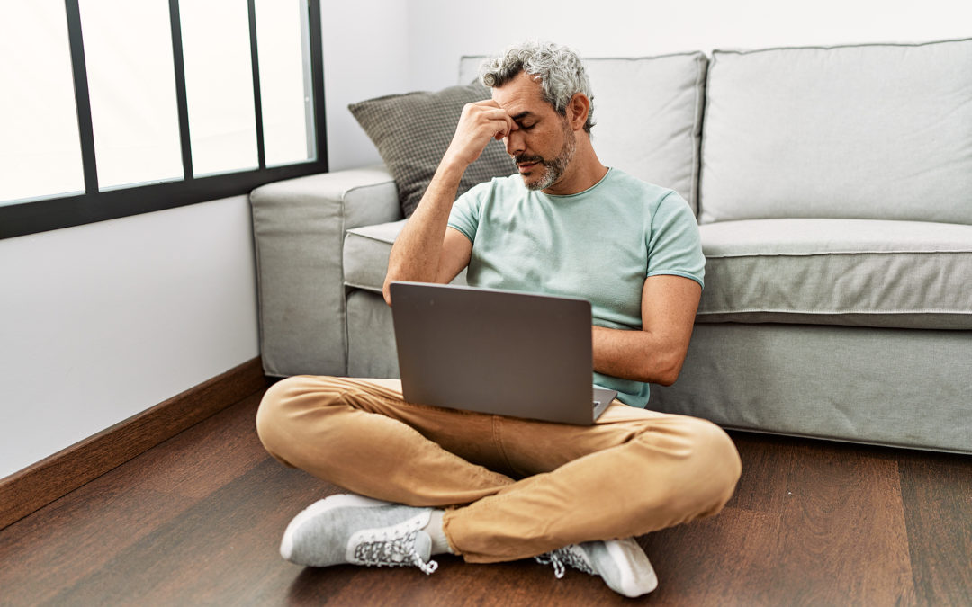 Man sitting on the floor with a laptop, looking stressed or having a headache.