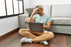 Man sitting on the floor with a laptop, looking stressed or having a headache.