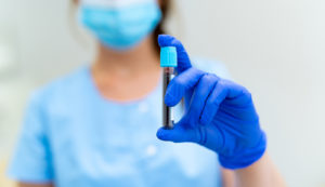 Healthcare professional in a mask holding a blood sample tube while wearing blue gloves.